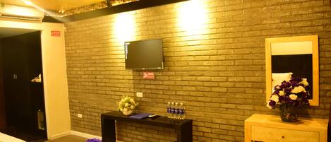 Ein Hotelzimmer mit Backsteinwand, einem flachbildfernseher über einer Holztruhe und einem Bett mit roter Bettwäsche, sowie einem kleinen Holztischchen mit Blumenvase.