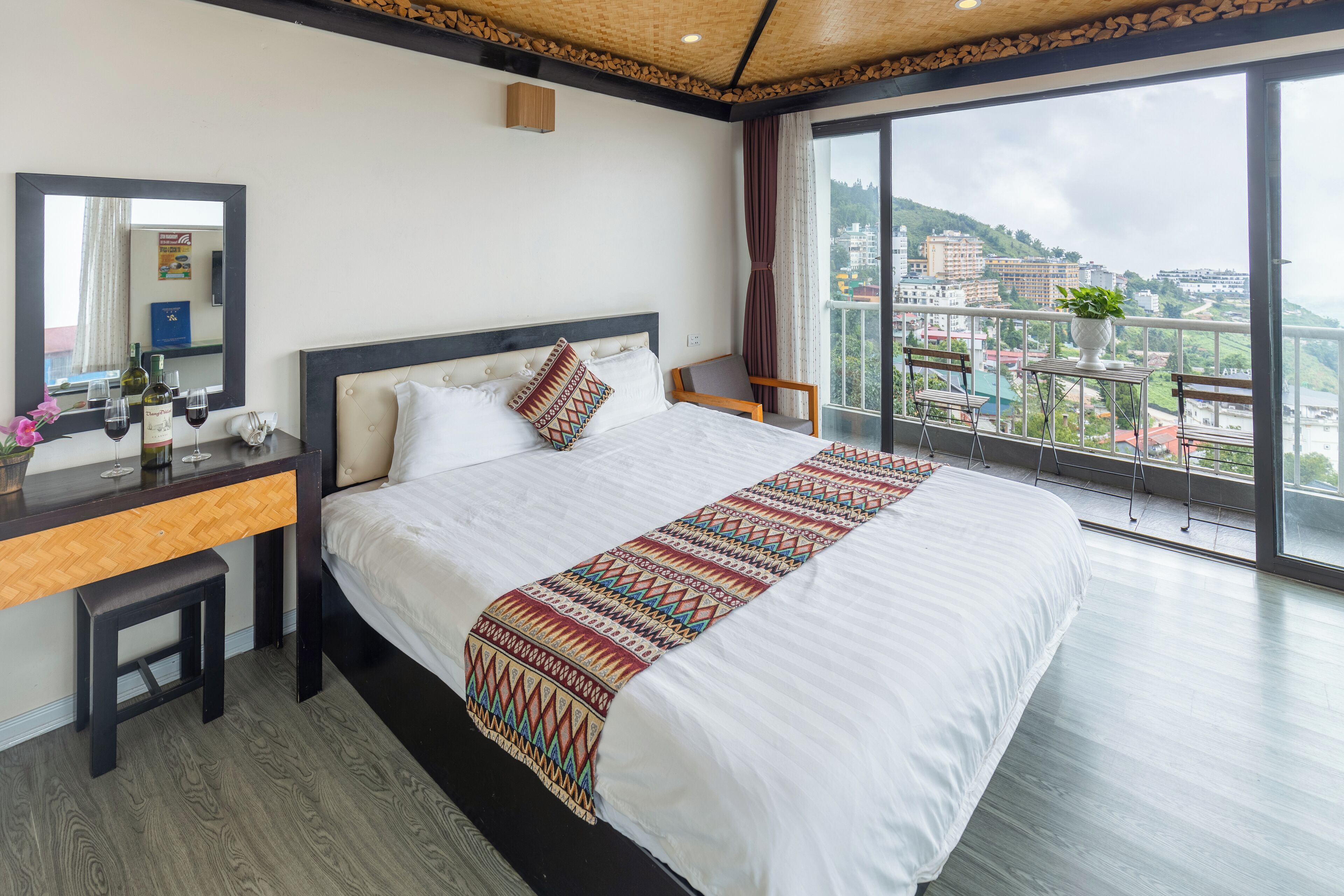 Habitación de hotel con una cama grande, un escritorio de madera, un espejo y un balcón con vista a la ciudad.