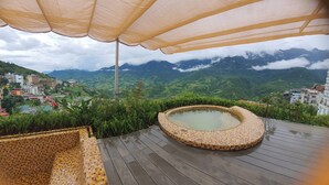 Een hottub op een houten terras met uitzicht op een bergachtig landschap en gebouwen op de achtergrond.