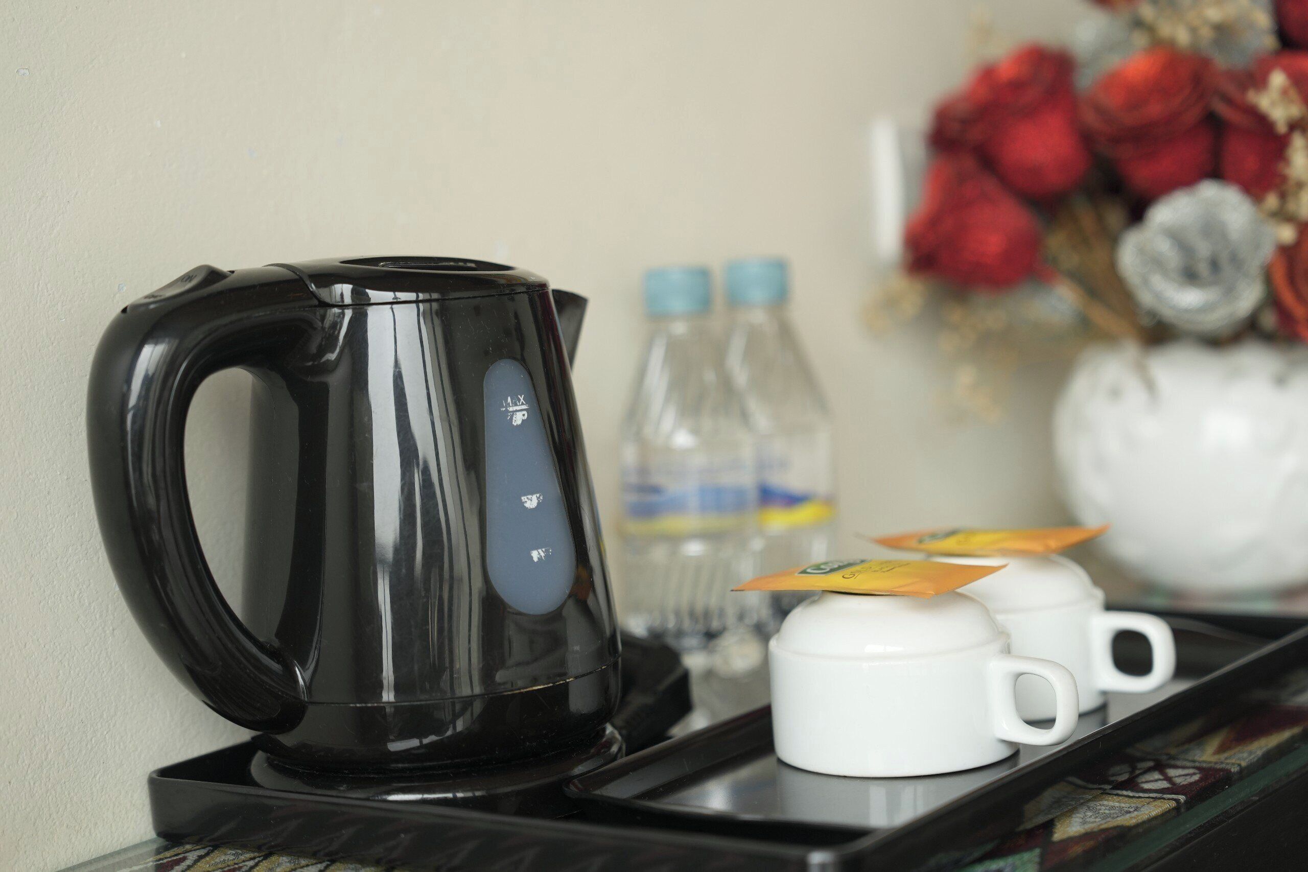 Una tetera eléctrica negra sobre una bandeja con dos tazas blancas y una tetera, una botella de agua, y un ramo de rosas rojas al fondo.