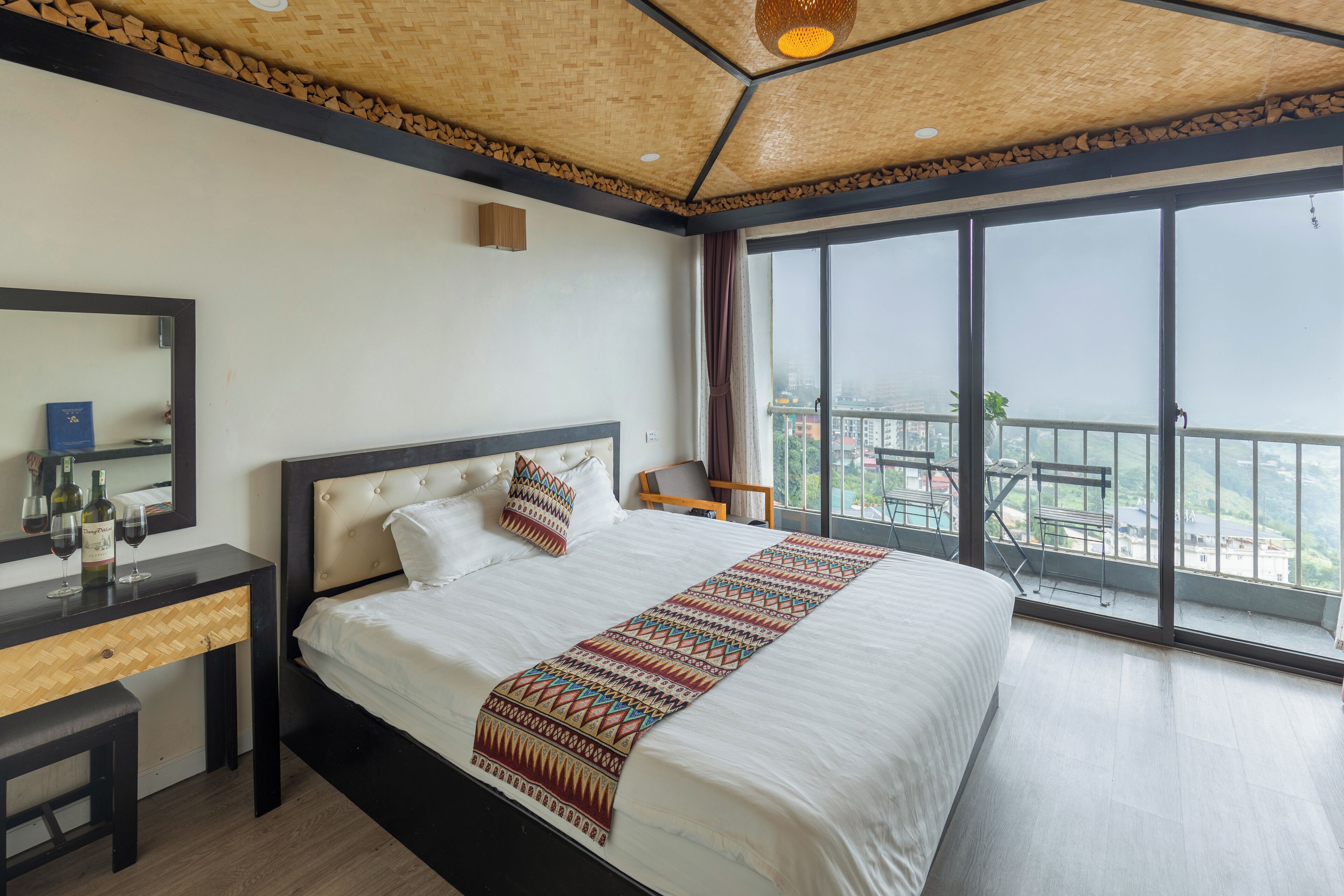 Ein Hotelzimmer mit einem großen Bett, einem Holztisch, einem Spiegel und Blick auf die Stadt durch große Fenster.