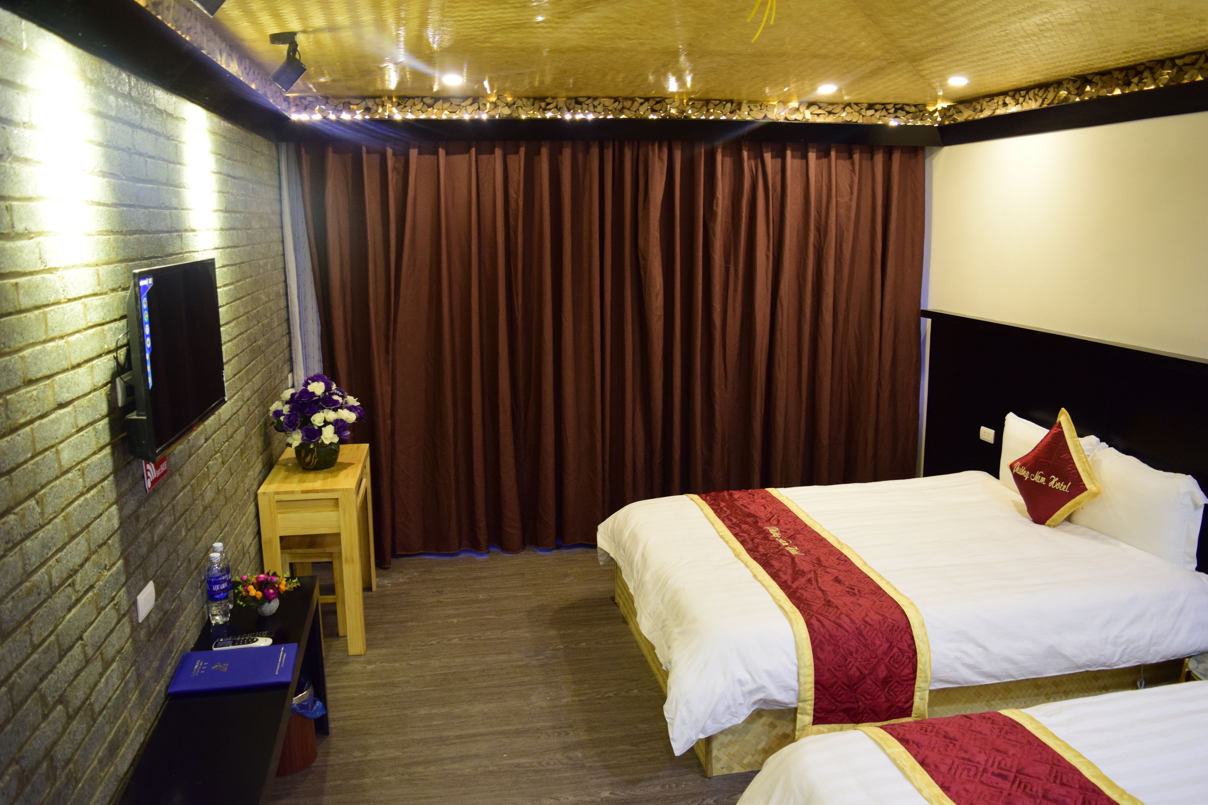 A hotel room with two beds, a TV on the wall, a small table with a vase of flowers, and a curtain.