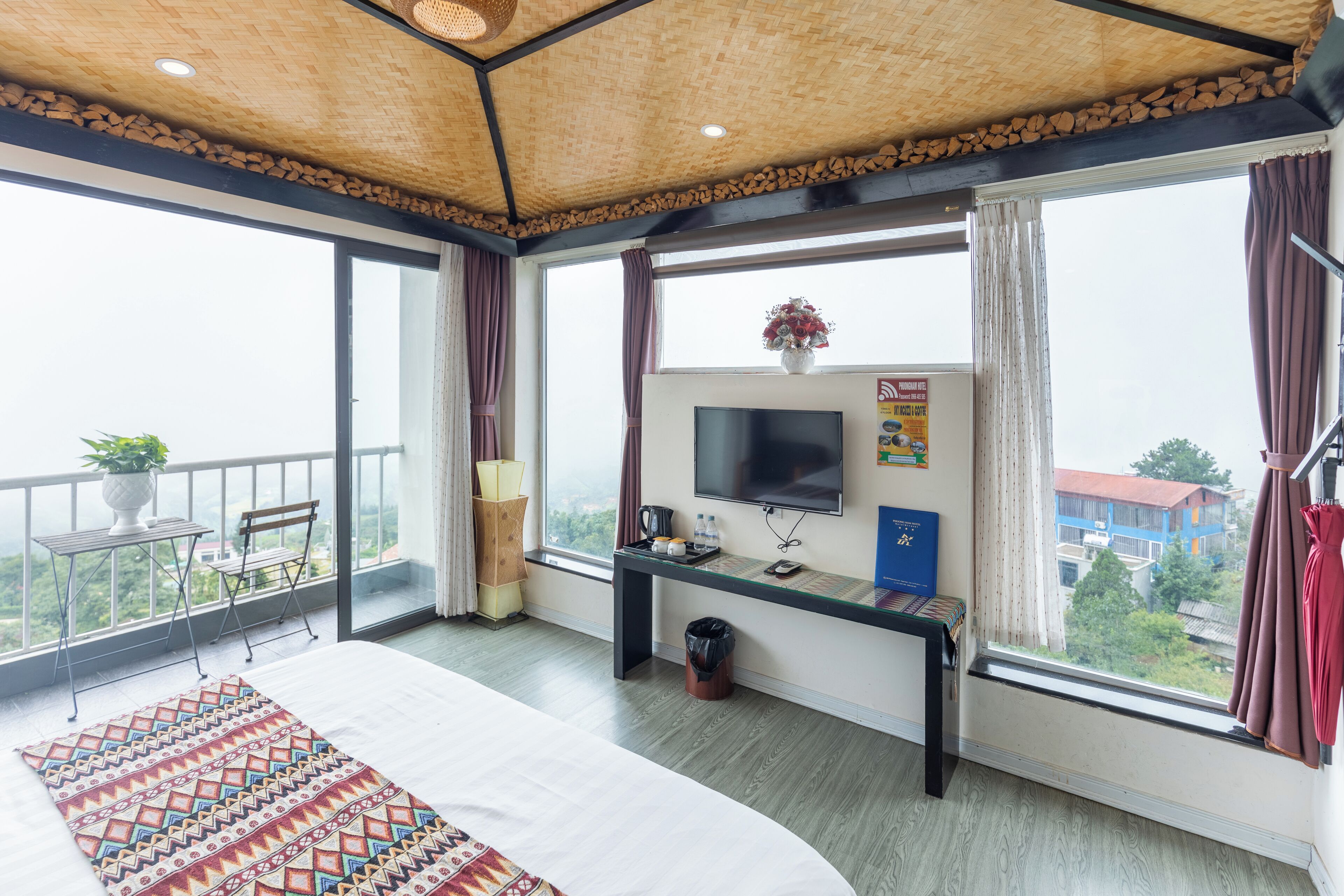 A hotel room with a bed, a desk with a TV, a window with curtains, a balcony with a table and chairs, and a view of buildings outside.