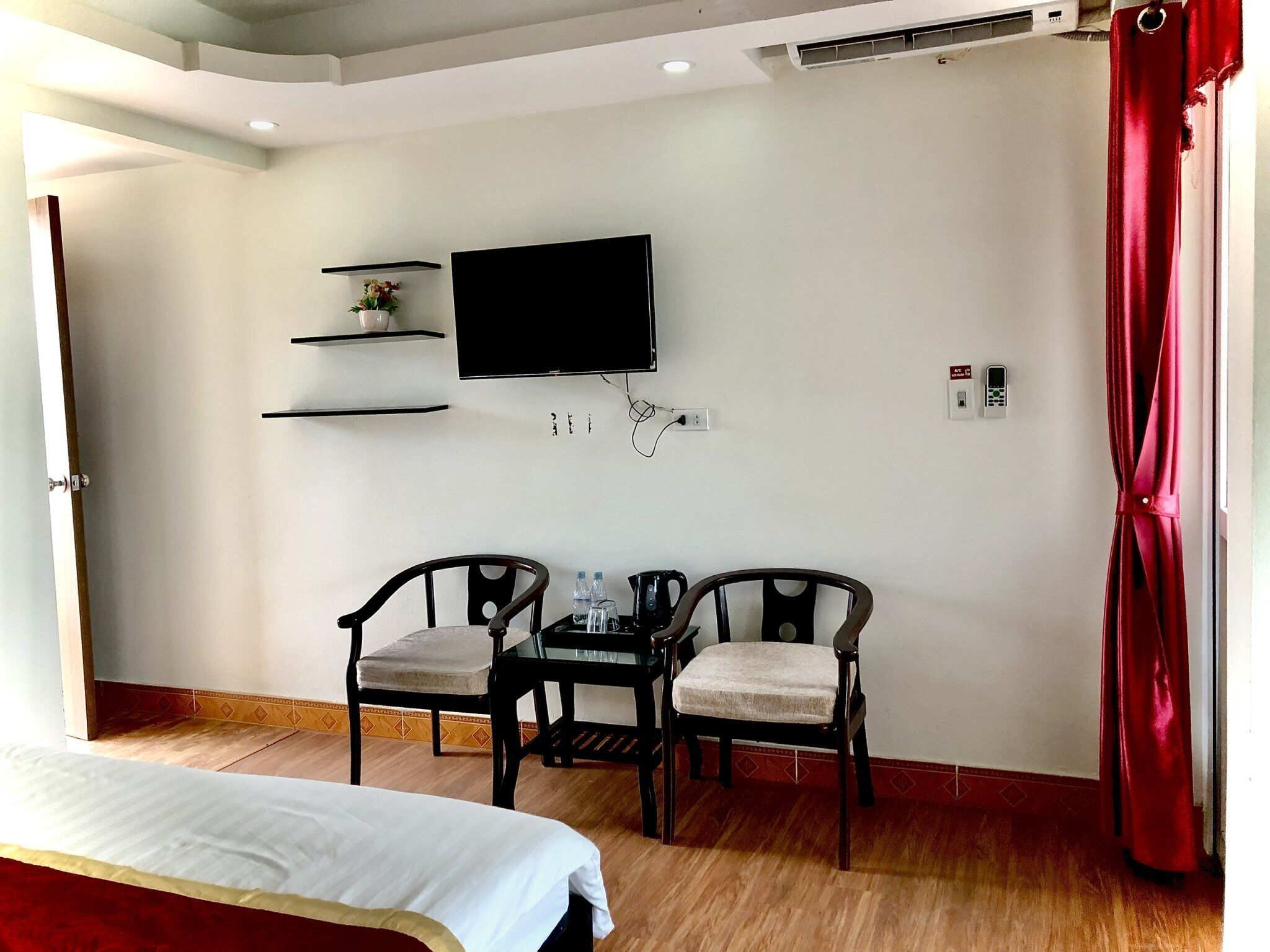 A hotel room with a bed, a desk with two chairs, a TV mounted on the wall, and a red curtain.
