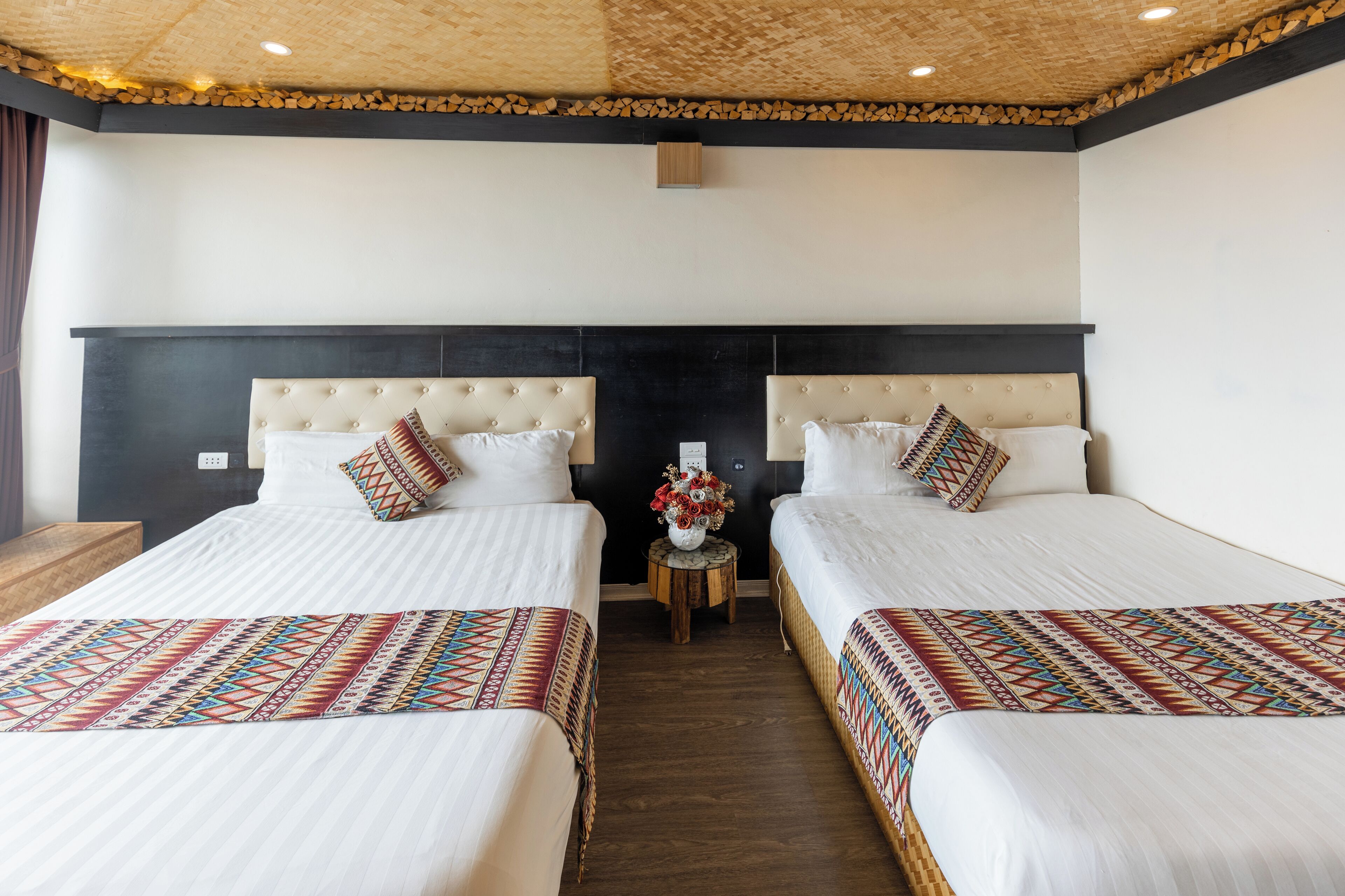 Habitación de hotel con dos camas, cabecero de madera, una mesita redonda con un florero y un techo con diseño tejido.
