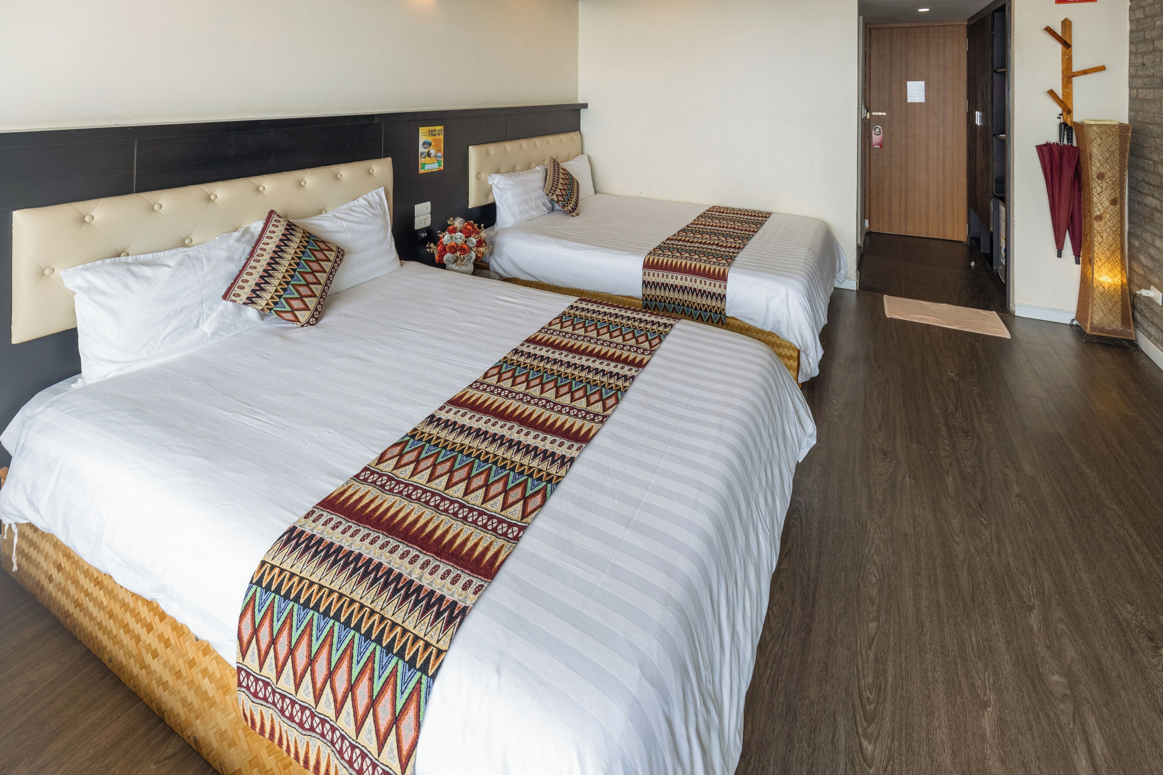 A hotel room with two beds, each with a patterned bedspread and pillows. The room has a wooden floor and a dark headboard.