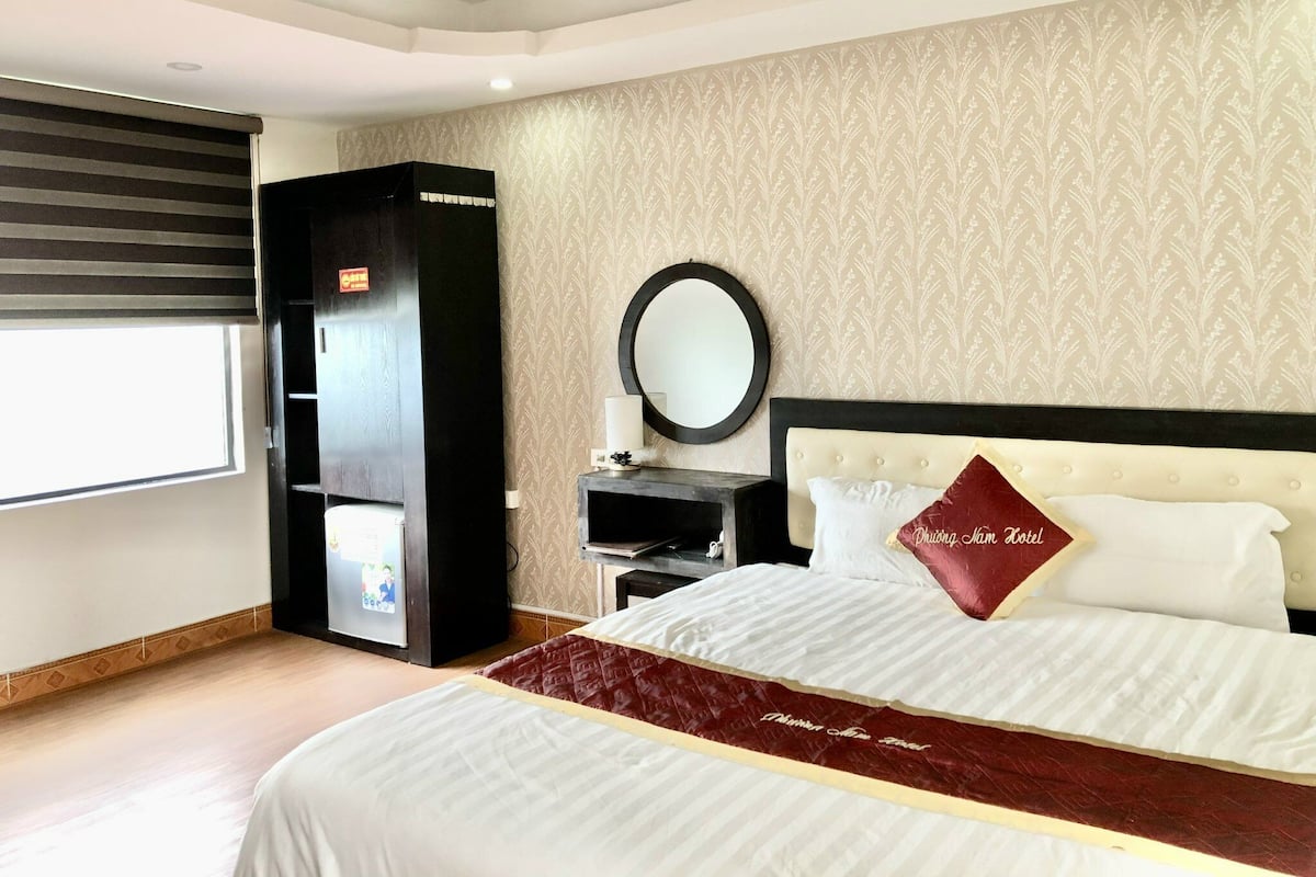Een hotelkamer met een groot bed, een garderobe, een ronde spiegel en een raam met jaloezieën.