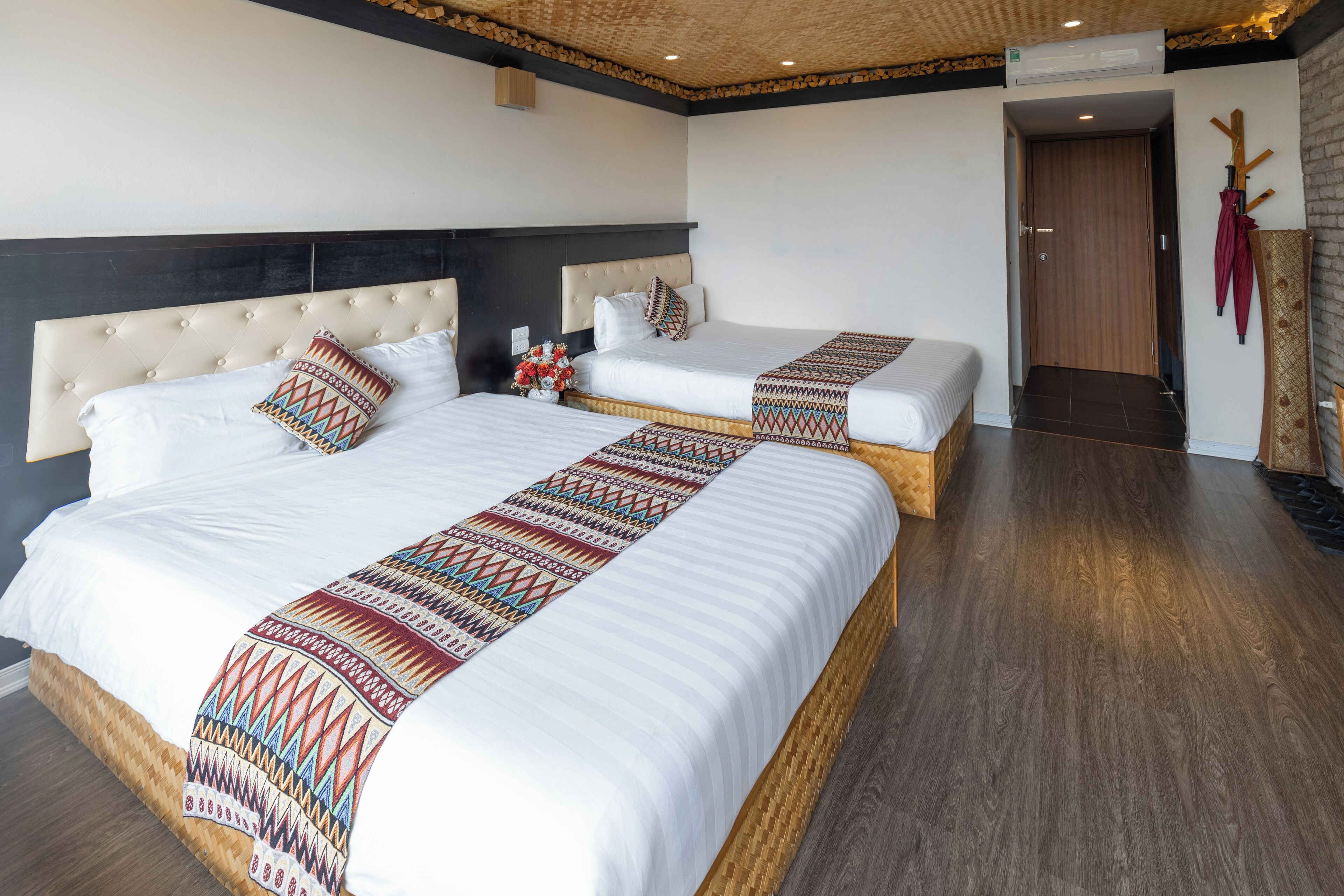 Quarto de hotel com duas camas, piso de madeira, teto com padrão trançado e porta com moldura de madeira.