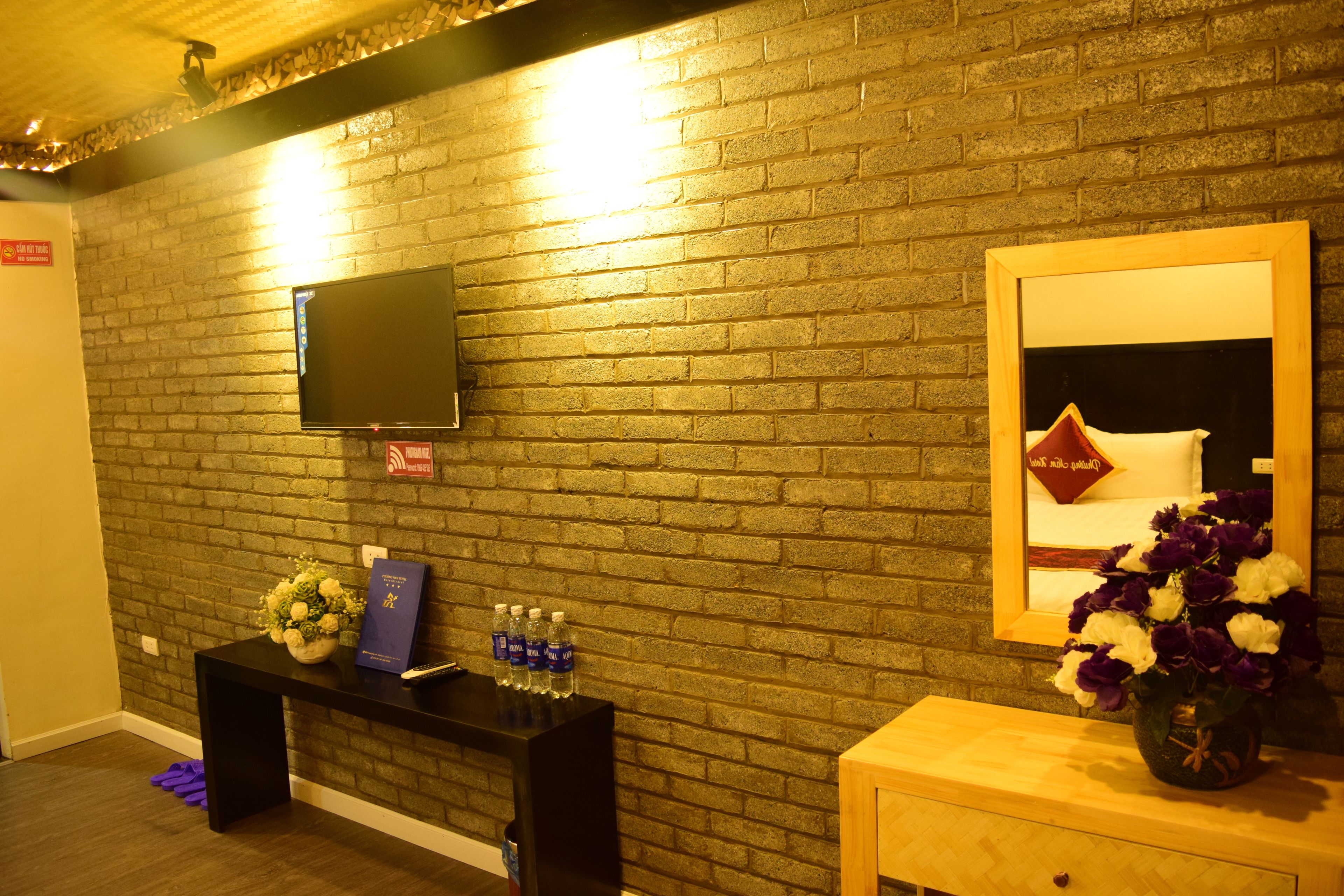 A hotel room with a brick wall, a flat-screen TV mounted above a black console table, a wooden bedside table with a vase of flowers, and a mirror reflecting a pillow and a red heart-shaped decoration.