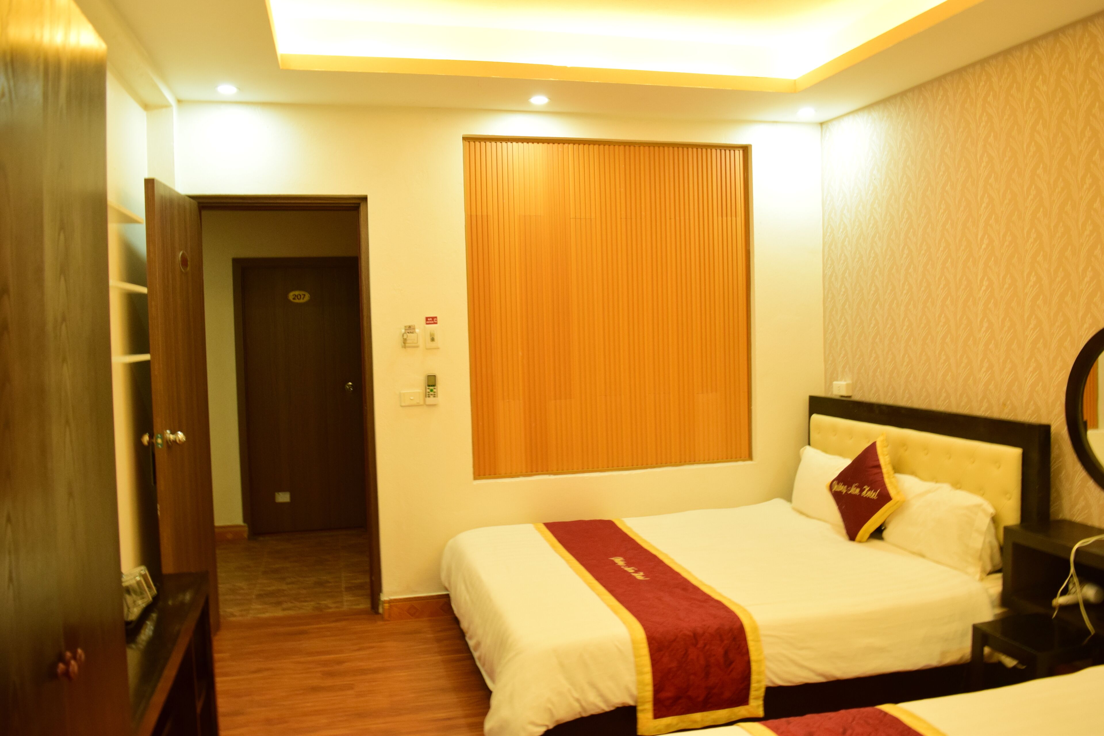 A hotel room with two beds, a wooden door, a wall-mounted light, and a small bedside table.