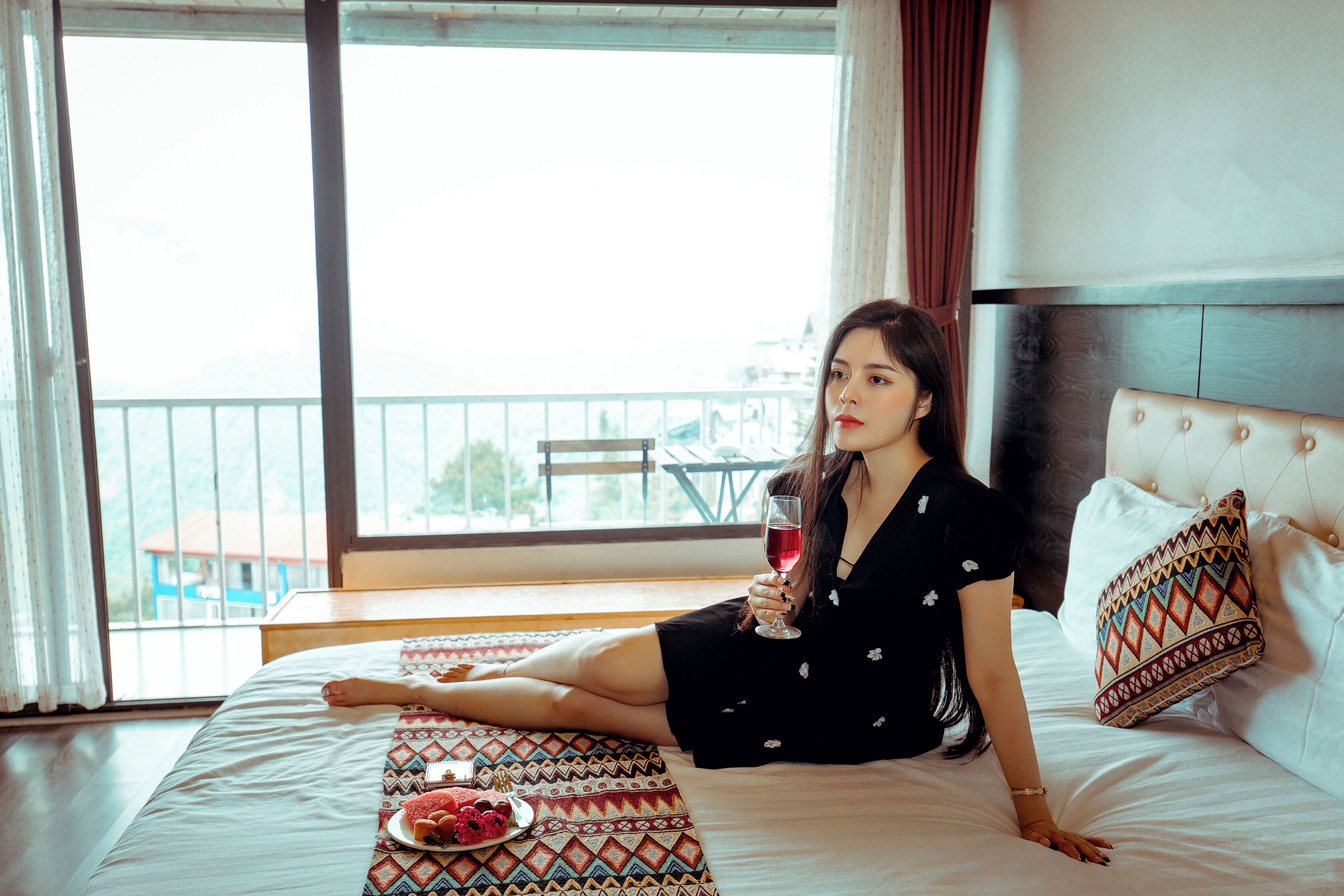 A woman sitting on a bed in a hotel room with a balcony view.