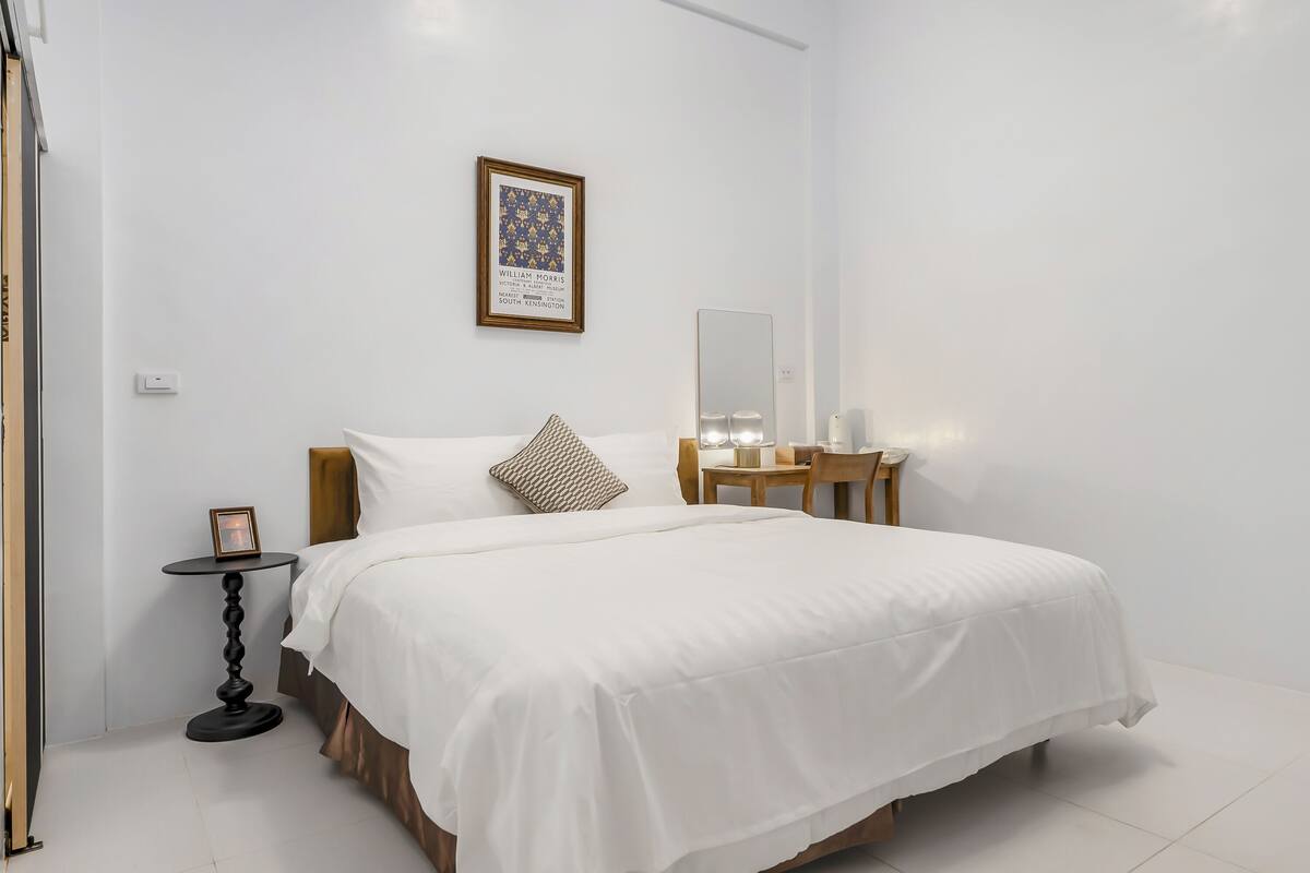 A neatly arranged bedroom with a large bed, a bedside table, a small round table, a mirror, and a framed picture on the wall.