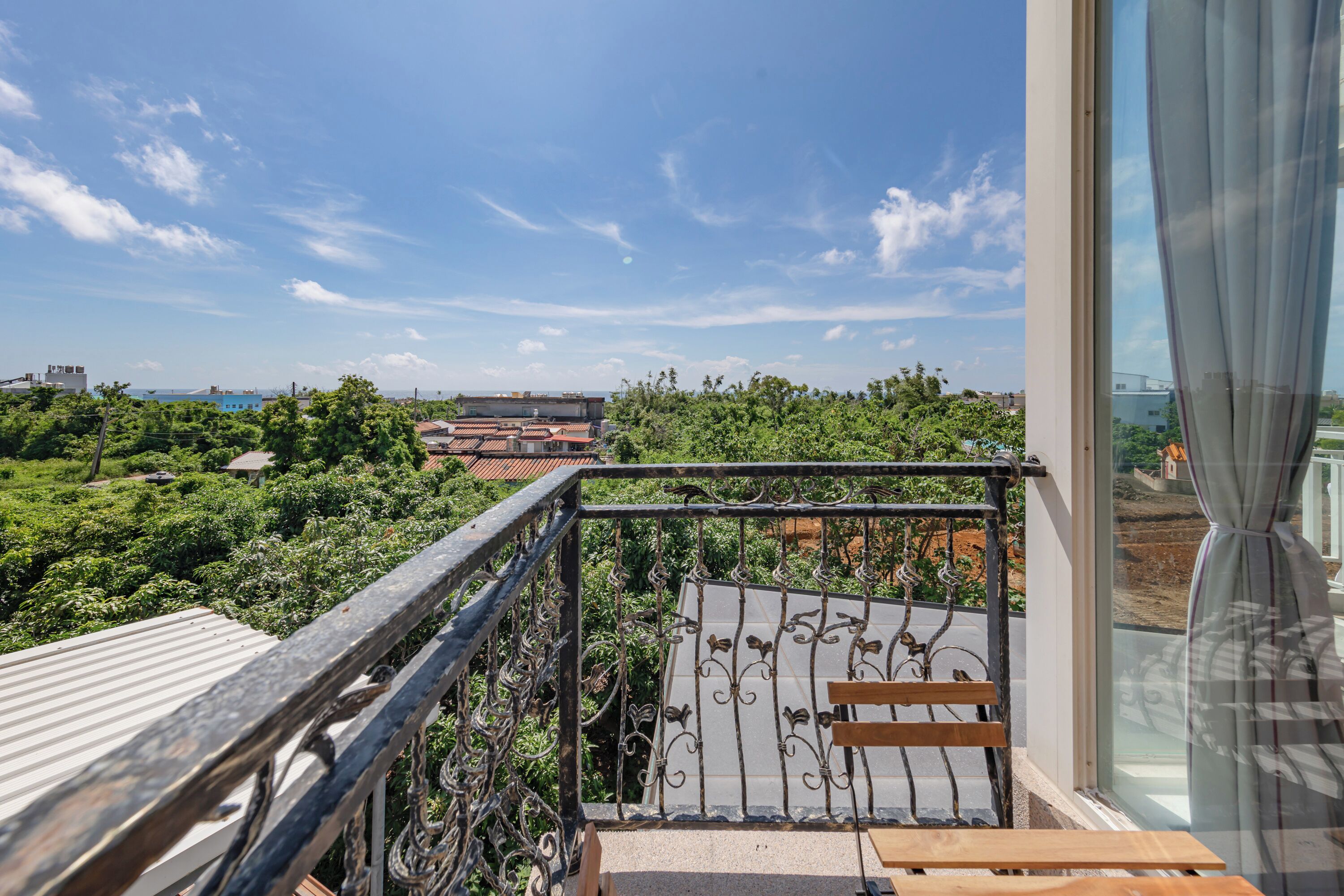 A balcony with a metal railing, a wooden bench, and a view of greenery and buildings in the distance.