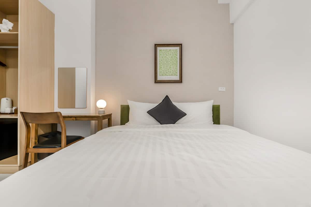 A hotel room with a bed, a desk, a chair, a bedside table, a lamp, and a framed picture on the wall.
