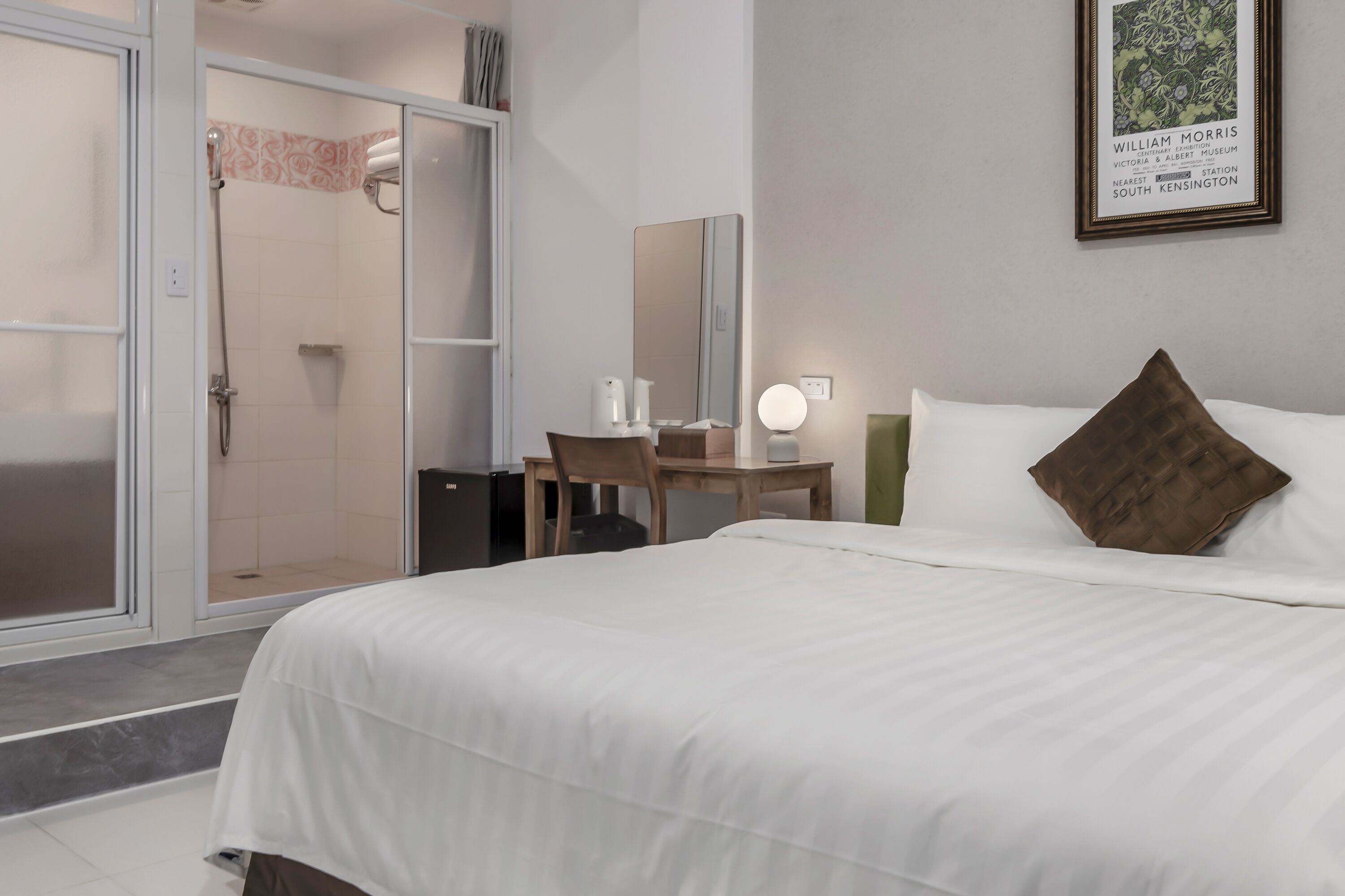 A hotel room with a bed, a desk with a chair, a shower, and a framed picture on the wall.