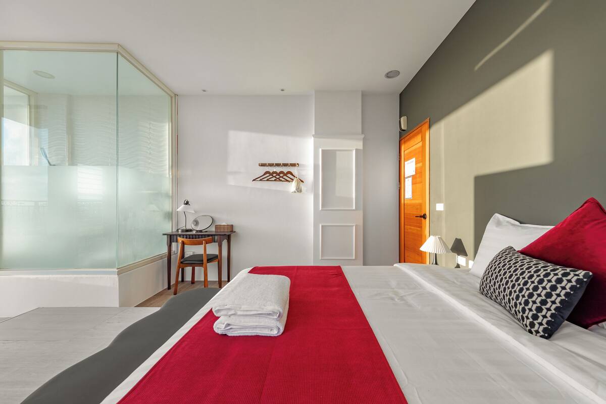 A hotel room with a large bed, a red and white bedspread, a glass shower enclosure, a wooden chair, a small table with a lamp, and a coat rack.