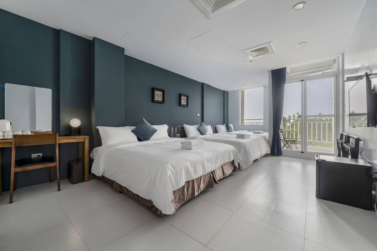 A modern hotel room with two beds, a desk, and a balcony with a view.