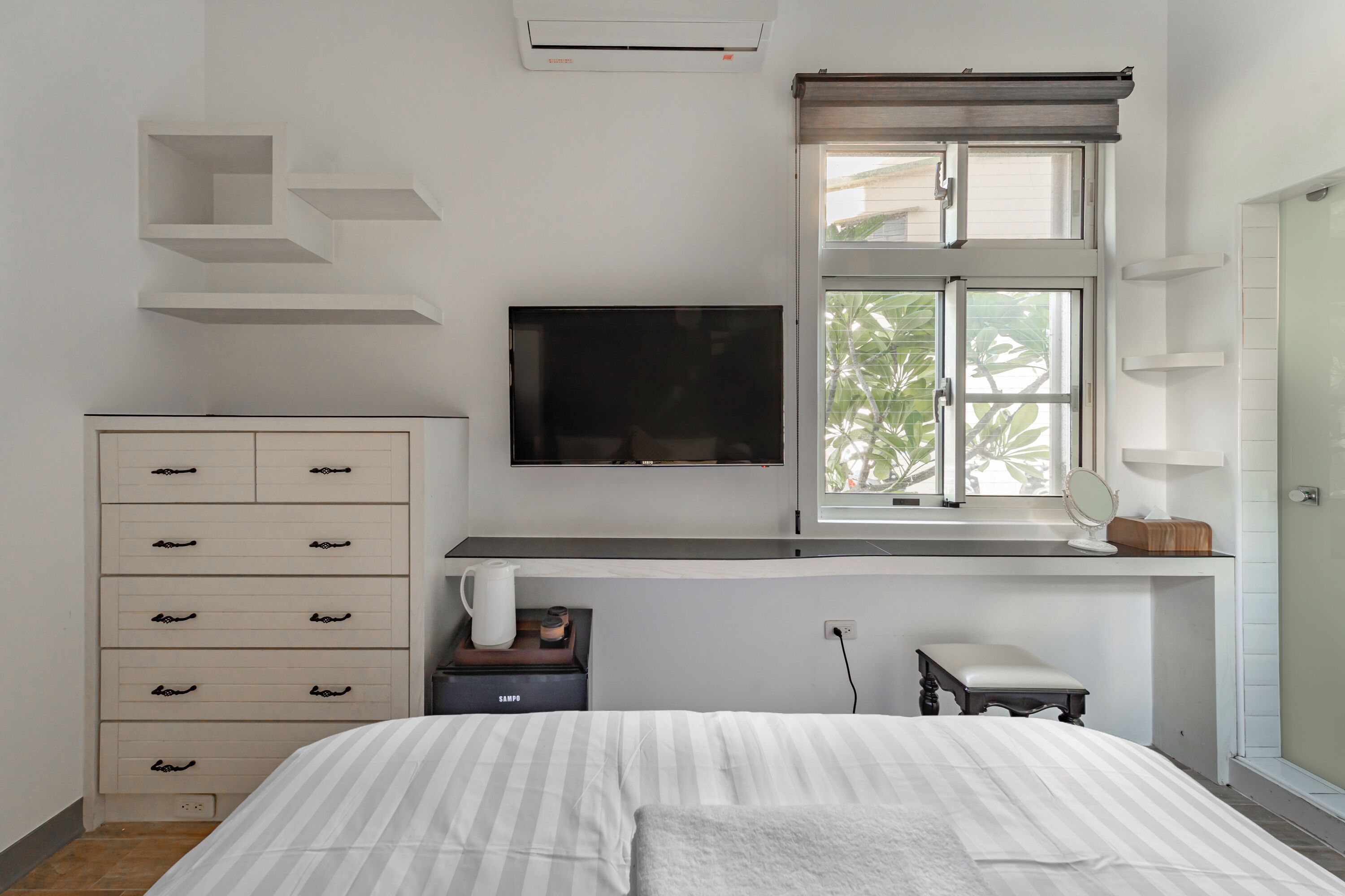 A bedroom with a bed, a TV mounted on the wall, a dresser, a window with blinds, and a small table with a lamp.