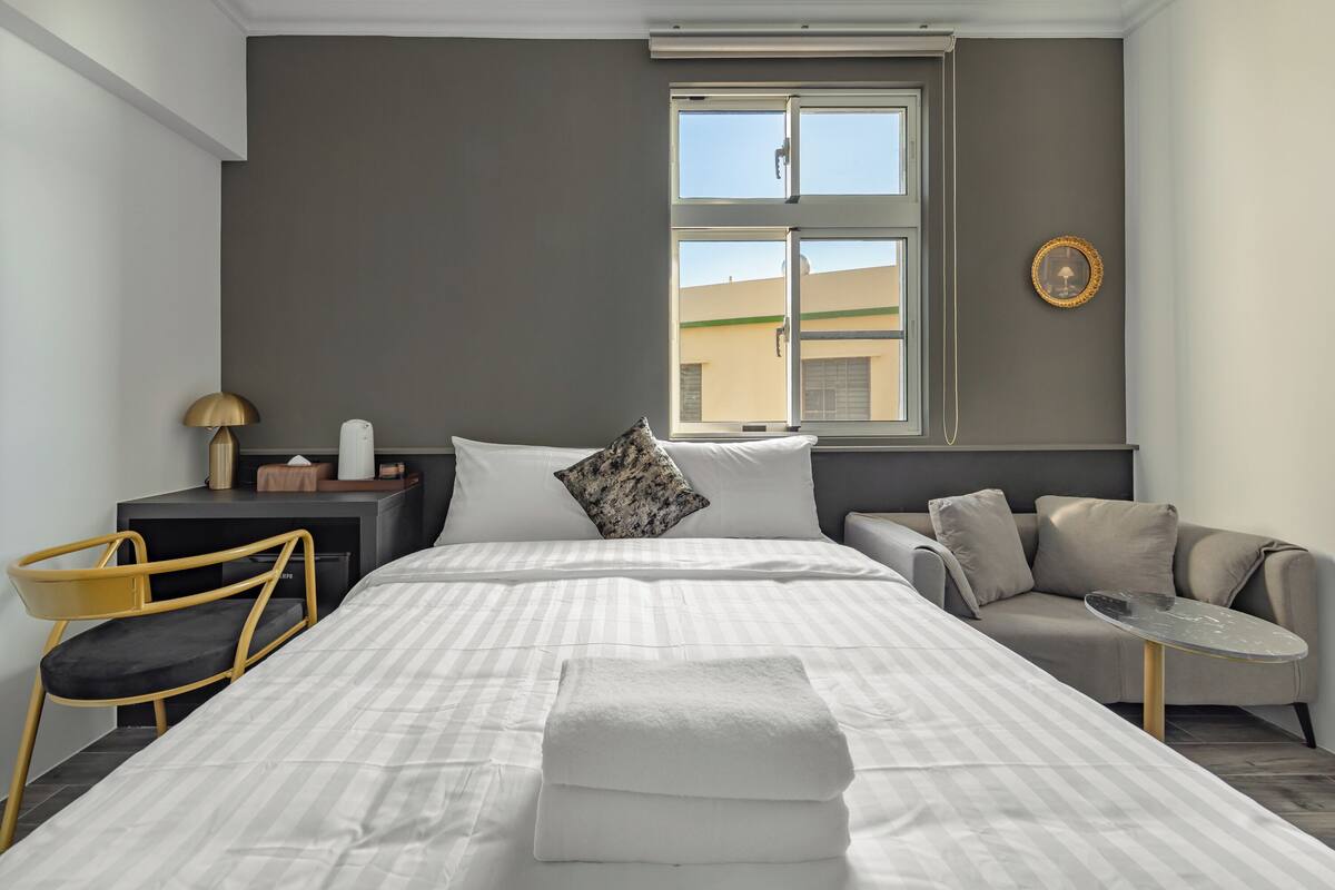 A modern hotel room with a large bed, a sofa, a small table, and a desk with a lamp.