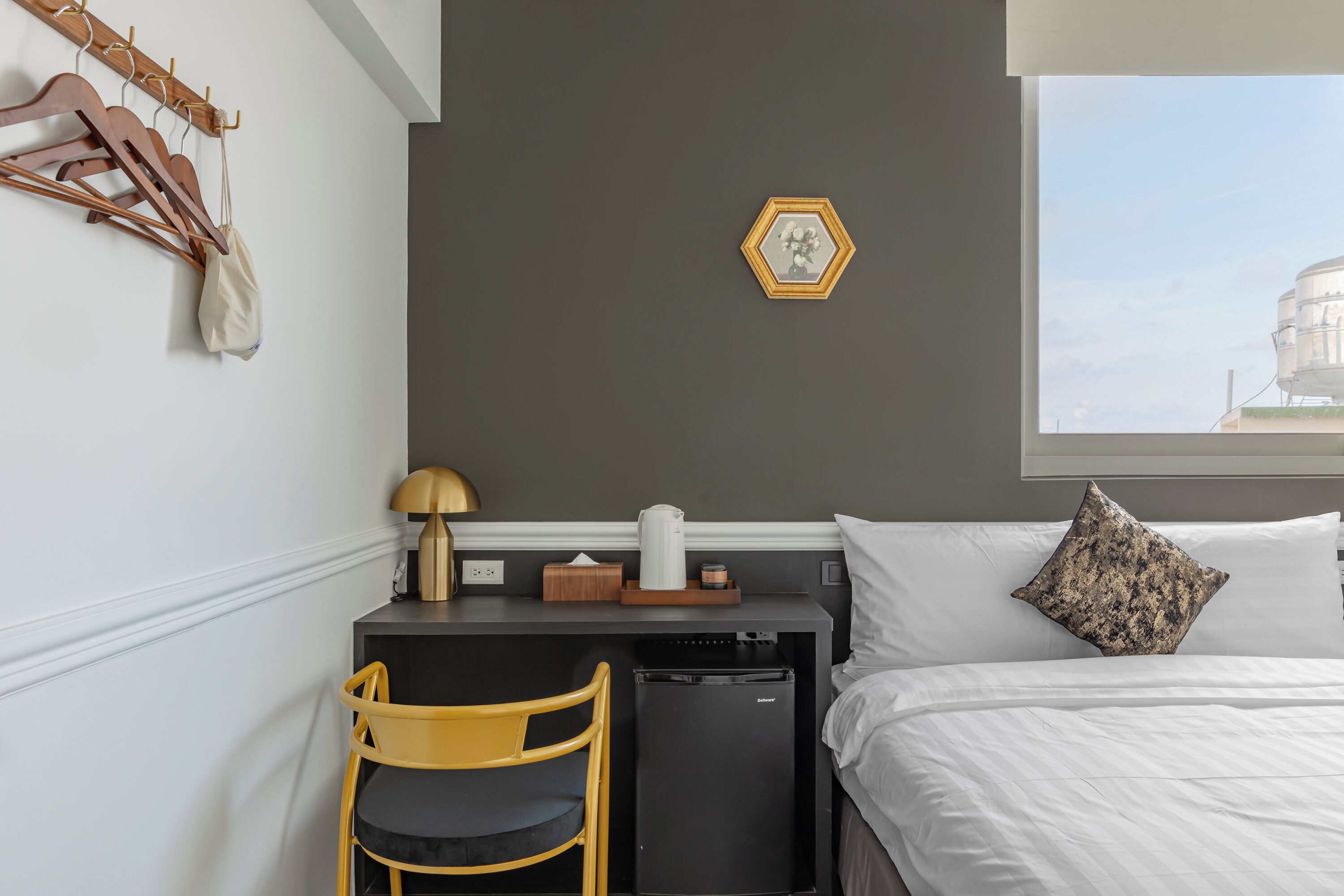 Ett hotellrum med en säng, ett skrivbord med en stol, ett litet kylskåp, en lampa, ett fönster med utsikt över en byggnad och en klädställning med galgar.