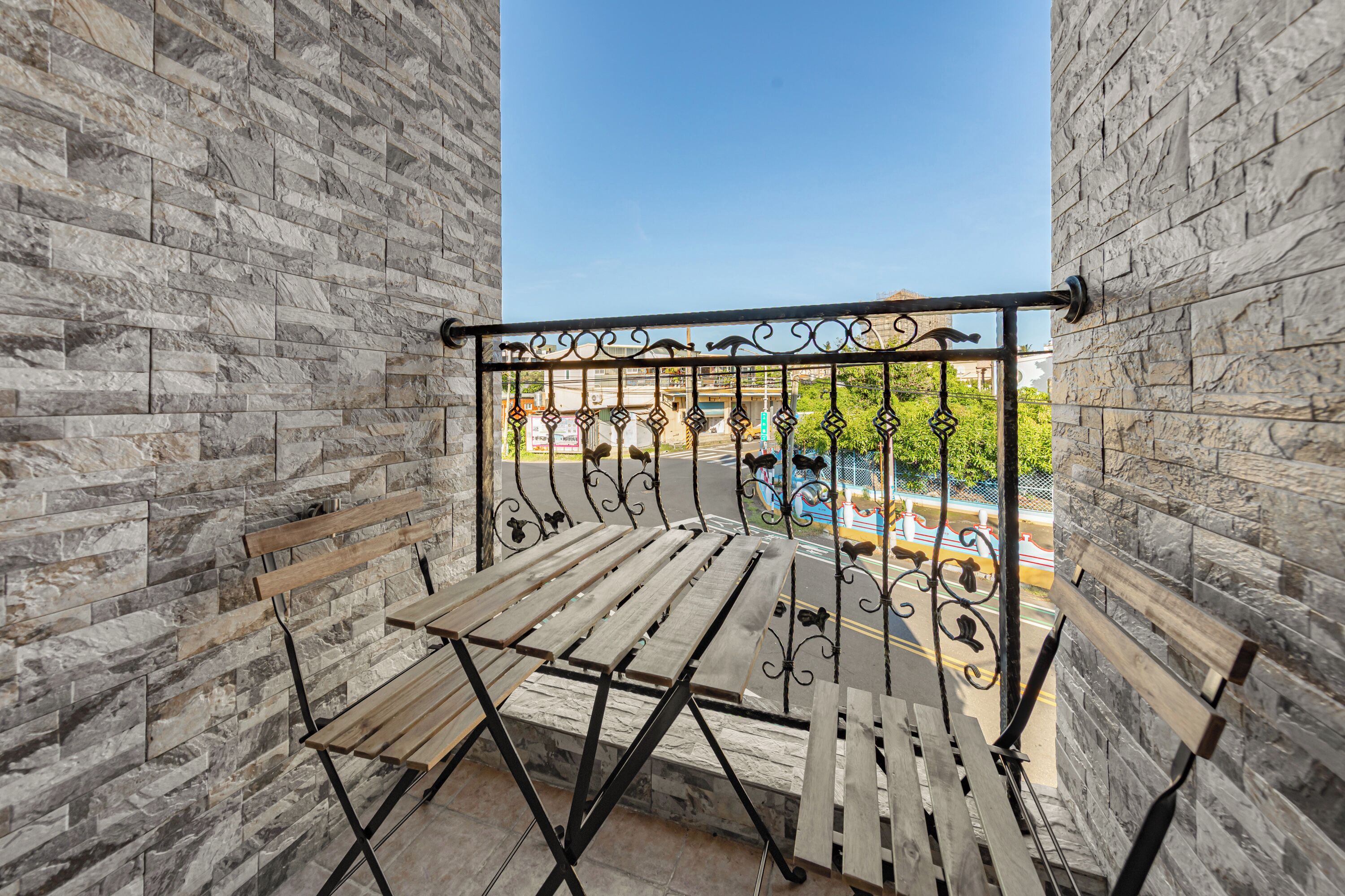 A balcony with a wrought iron railing, wooden benches, and a view of a street with shops and greenery.