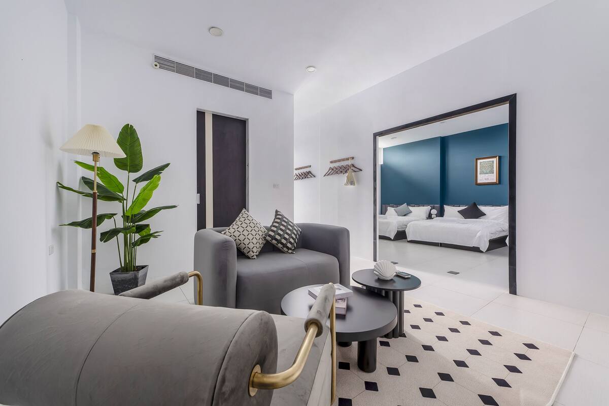 A modern hotel room with a grey sofa, a small round table, a potted plant, and a large mirror reflecting the room.