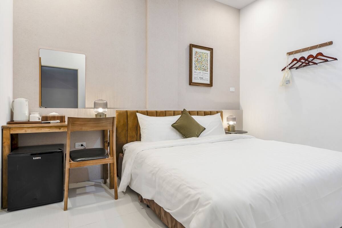 A hotel room with a bed, a desk with a chair, a small refrigerator, a mirror, a framed picture on the wall, and a coat rack with hangers.