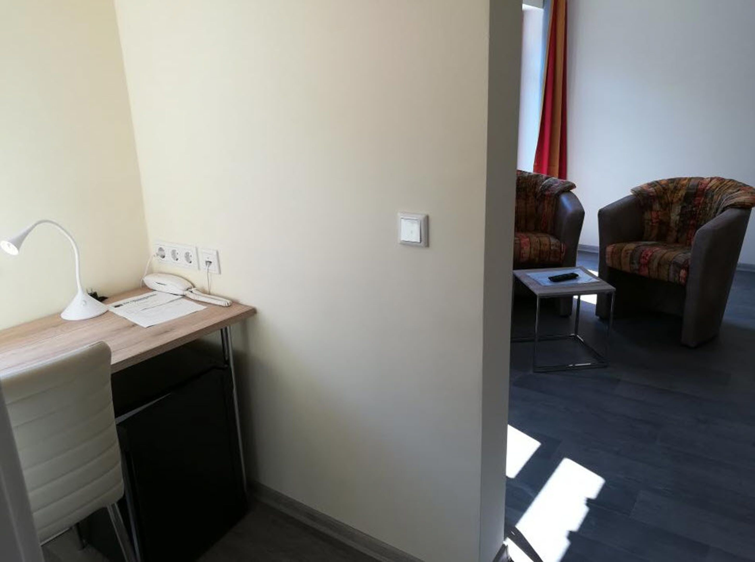 Una habitación pequeña y bien iluminada con un escritorio, una silla y un sofá. Tiene una ventana con cortinas, y el diseño es sencillo y limpio.