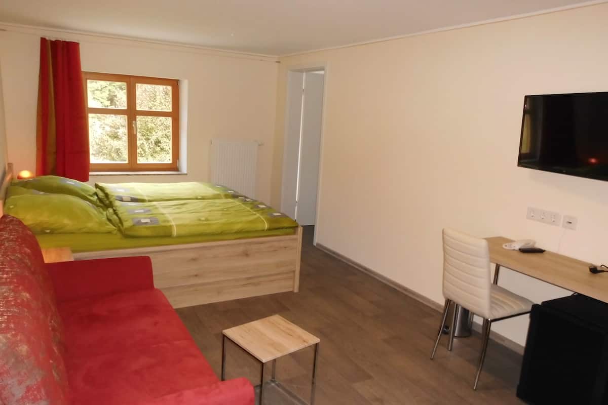 Ein Zimmer mit einem Bett, einem roten Schlafsofa, einem kleinen Holztisch, einem Schreibtisch mit Stuhl und einem an der Wand befestigten Fernseher.