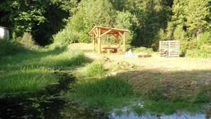 Un kiosque en bois, situé dans une zone herbeuse, surplombe un étang dans lequel se reflètent les arbres.
