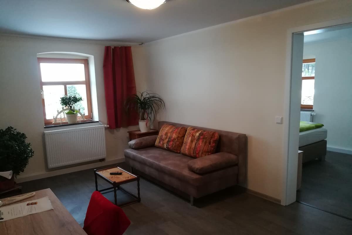 Ein Wohnzimmer mit braunem Sofa, einem kleinen Tisch, einem Sessel, einer Heizung, einem Fenster mit roten Vorhängen und einer Zimmerpflanze.