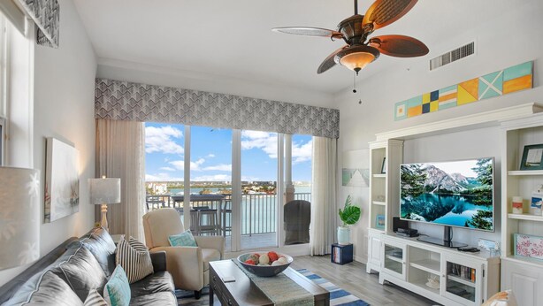 A modern living room with a sofa, coffee table, and TV. There is a balcony with a view of the city and a ceiling fan.