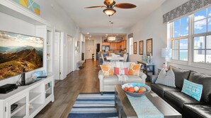A spacious living area with a large flat-screen TV, a ceiling fan, and a view of the kitchen and dining area.
