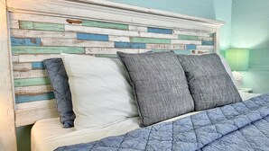 Una cama con colcha azul acolchada, almohadas blancas y almohadas grises, y un cabecero con diseño de madera pintada.