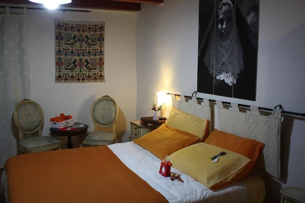 A room with a bed, two chairs, a small table with a fruit basket, a wall tapestry, and a framed picture.