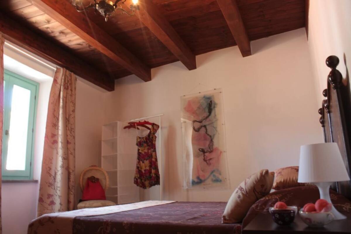 A bedroom with a wooden ceiling, a bed with a patterned bedspread, a nightstand with a lamp, and two abstract paintings on the wall.