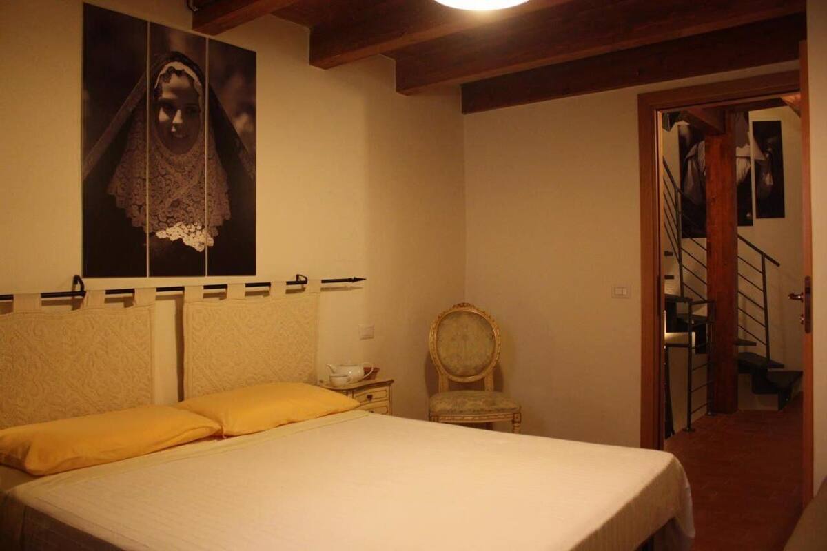 A bedroom with a bed, a chair, a poster on the wall, and a staircase in the background.