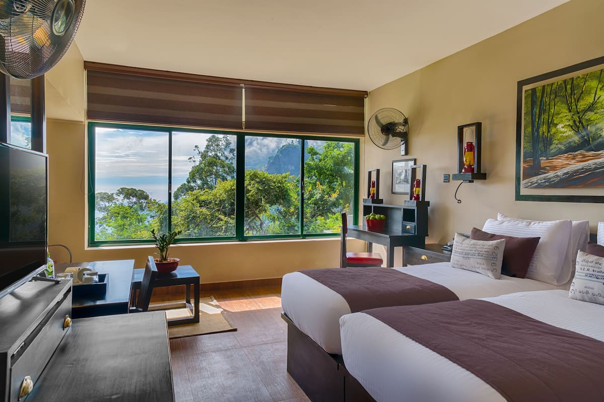 A hotel room with a large window, a bed, a desk, a chair, a TV, and a fan.