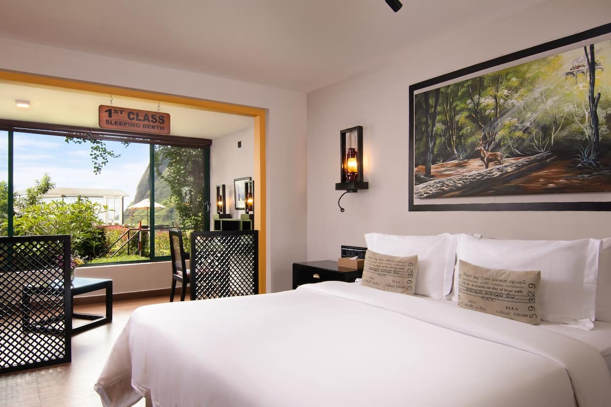 A bedroom with a bed, a nightstand, a wall-mounted lamp, a painting, and a window with a view of greenery and a building outside.
