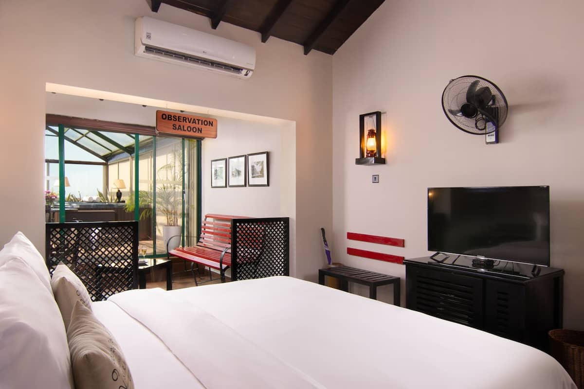A hotel room with a bed, a TV, a desk, and a chair. There's a sign that says "Observation Saloon" and a fan on the wall.