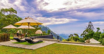 Un patio con sillones de descanso y sombrillas, rodeado de exuberante follaje y un paisaje montañoso.