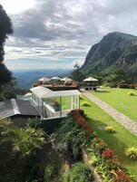 Un edificio moderno con cerramiento de vidrio, rodeado de exuberante vegetación y un telón de fondo montañoso.