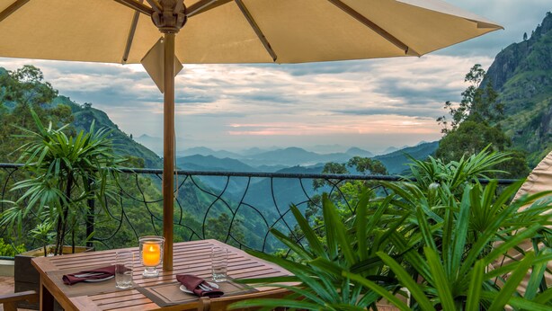 A table with a table umbrella, a candle, and a glass of water, overlooking a mountainous landscape. - EKHO Ella (Ella)
