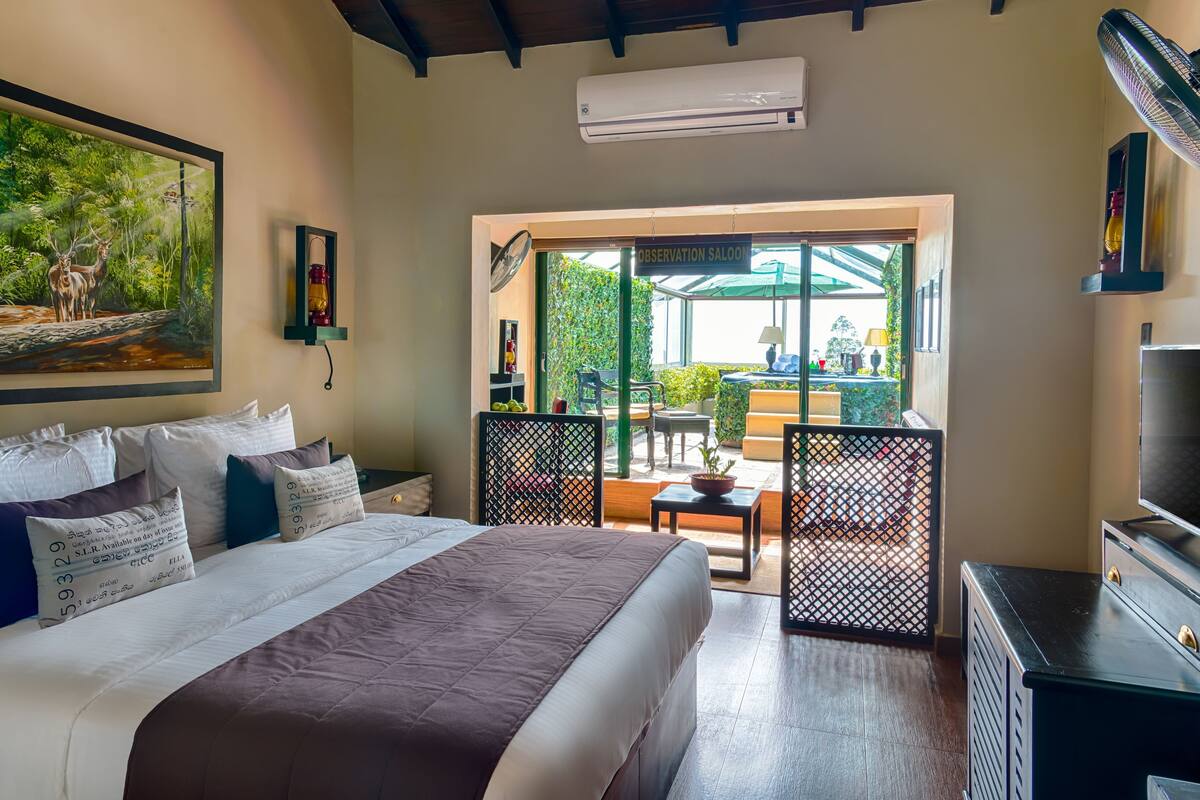 A bedroom with a bed, pillows, a painting, an air conditioner, a TV, and a view of the outside.