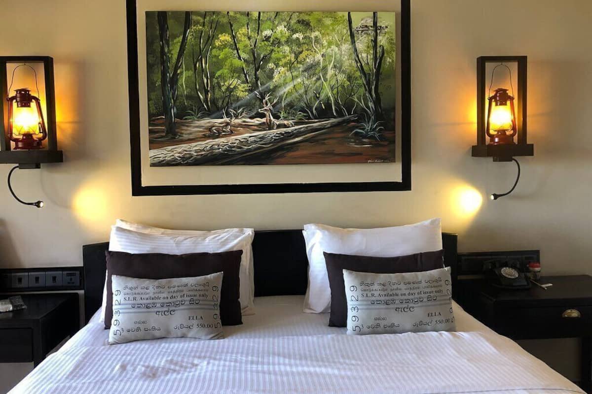 A hotel room with a bed, two pillows, a painting of a forest scene, and two wall-mounted lanterns.