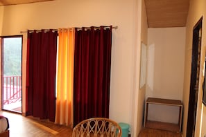 A room with a wooden floor, a wicker chair, a small table, and a window with red curtains. - Beyond Stay Kastura Kanatal (Dhanaulti)