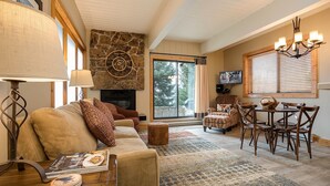 A cozy living room with a stone fireplace, a dining area with wooden chairs, and a large window with blinds. - CA111 | Pool | Slopeside w/ Great Views | Winter Shuttle | Hot Tub (Steamboat Springs)