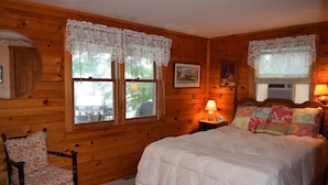 A wooden cabin bedroom with a bed, a chair, and two windows with lace curtains. - Three month rental minimum.   (Greenland)