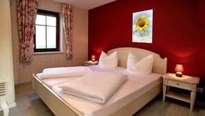 A neatly made bed with white linens, two bedside tables with lamps, a window with floral curtains, and a sunflower painting on the wall. - Villa Rosamunde (Burg (Spreewald))