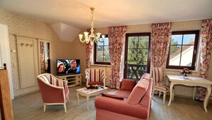 A living room with a sofa, armchair, TV, and floral curtains. - Villa Rosamunde (Burg (Spreewald))